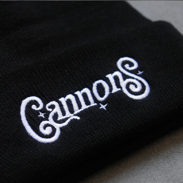 close up image of a black winter beanie on a concrete floor. front cuff of beanie has white embroidery that says cannons with three stars