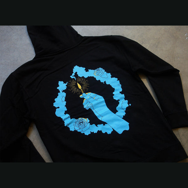  Image of the back of a black hooded sweatshirt on a white background.  The back of the hoodie is blue and features a hand holding up a match with a yellow flame and an aura around the match. Surrounding the hand is a circle flower wreath in blue.