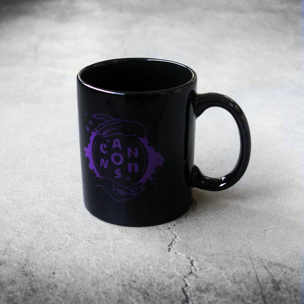 image of a black coffee mug on a concrete floor. purple print on the front of the mug of two hards holding a crystal ball and the word CANNONS inside of the ball