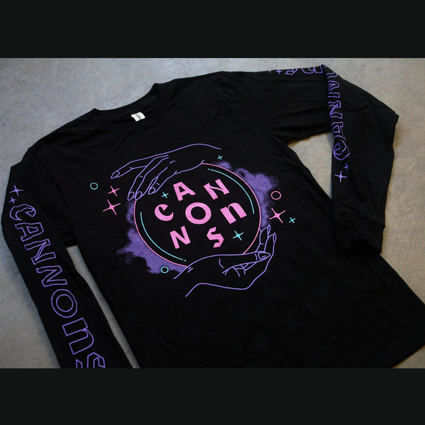 Close up photo of a black longsleeve on a gray background. The front of the long-sleeve features two purple hands on a crystal ball, with pink letters spelling out the word cannons. There are pink and green stars and circles around the design and a purple cloud aura behind and around the crystal ball. On the sleeves in purple there are stars and it says Cannons in outline writing with no fill.